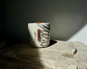 Pode incluir: Uma vela de cerâmica branca com design marmorizado preto e pavio de madeira. A vela tem um rótulo retangular com a palavra "malmö" impressa. A vela está sobre uma superfície de pedra cinza.