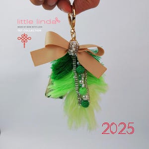 Könnte beinhalten: Ein grüner und hellgrüner Quasten-Schlüsselanhänger mit einer beige Schleife, Perlen- und grünen Perlenakzenten und einem goldenen Schlüsselring. Der Schlüsselanhänger trägt den Text "little linda" und "TET COLLECTION". Das Jahr 2025 ist in Rot unter dem Schlüsselanhänger geschrieben.