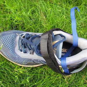 Può includere: Una scarpa sportiva blu e grigia con lacci blu navy e un cinturino nero. Un cinturino blu è attaccato alla scarpa. La scarpa è sull'erba verde.