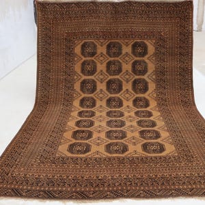 Puede incluir: Una alfombra rectangular marrón con un patrón central de octágonos y diamantes repetidos. La alfombra tiene un borde detallado con diseños geométricos. La paleta de colores incluye tonos marrones y beige, creando una estética cálida. La alfombra se muestra sobre una superficie blanca.
