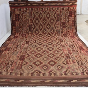 May include: A large, rectangular rug with a geometric diamond pattern in shades of brown, beige, and red. The rug has a detailed border and fringe along the edges. The design features repeating diamond and geometric shapes.