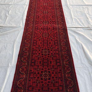May include: A long, narrow red rug with an intricate geometric pattern. The rug features a central field of red with black and gold accents, and a detailed border. The rug has a fringe at the end and is likely made of wool.
