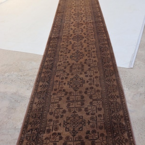 May include: A long, narrow runner rug featuring a deep brown base color with a repeating dark brown geometric and floral pattern. The rug has a distinct border design and short white fringe at the bottom edge, extending vertically across the floor.