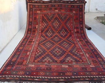 May include: A large, rectangular rug with a geometric diamond pattern in shades of burgundy, brown, and orange. The rug has a detailed border with floral and geometric motifs. Fringes are visible along the bottom edge.