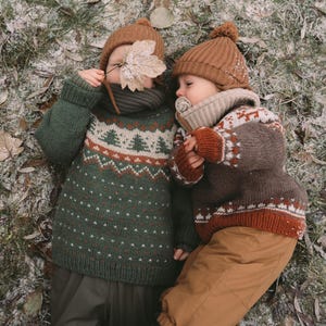 May include: Two children wearing knit sweaters and hats. One child wears a green sweater with a white and orange tree pattern and dark pants. The other child wears a brown sweater with a white and orange pattern, tan pants, and a beige pacifier.
