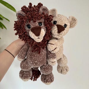 Op de afbeelding: Twee gehaakte leeuwen knuffels, een bruin en een beige, knuffelen elkaar. De bruine leeuw heeft een pluche manen.