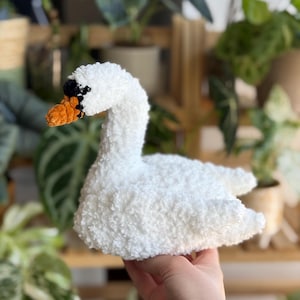 Könnte beinhalten: Ein handgefertigter weißer Plüschschwan mit strukturiertem Körper, einem orangefarbenen Schnabel und schwarzen Akzenten um das Auge. Der Schwan ist ein weiches, kuscheliges Spielzeug, perfekt für ein Kinderzimmer oder als Dekorationsartikel. Der Schwan wird vor einem Hintergrund aus grünen Pflanzen gehalten.