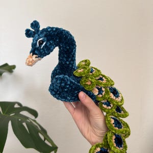 May include: A handmade peacock plushie in shades of teal, green, and tan. The plushie features a curved neck, a detailed head with a beak, and a fan of layered tail feathers. The soft, textured material gives the peacock a cuddly appearance.