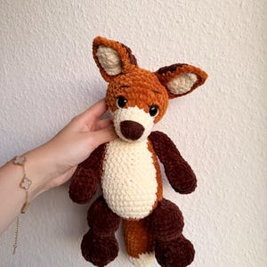 Könnte beinhalten: Ein handgefertigtes Fuchs-Plüschtier aus Häkelarbeit. Der Fuchs hat ein braunes und cremefarbenes Farbschema mit einem weißen Bauch und einem flauschigen Schwanz. Der Fuchs hat große, ausdrucksstarke Augen und ein niedliches, freundliches Gesicht. Das Spielzeug ist weich und kuschelig.