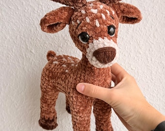 May include: A brown and white crocheted deer with large black eyes and a white muzzle. The deer has brown antlers and white spots on its back. The plush toy is held by a person.