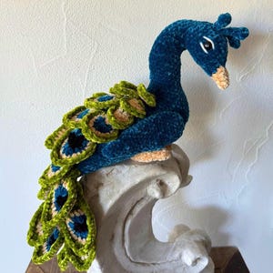 Könnte beinhalten: Ein gehäkeltes Plüsch-Pfauenspielzeug mit einem blaugrünen Körper und grünen und schwarzen Schwanzfedern. Der Pfau sitzt auf einem weißen, dekorativen Ständer.