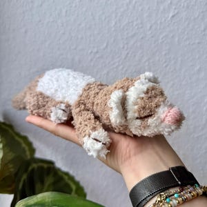 May include: A soft toy ferret in brown, white, and pink, lying on its side. The toy has a fluffy, textured appearance, with a white belly and face, and a pink nose. The ferret is a handmade item.