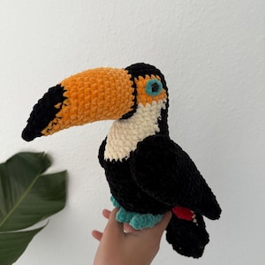 Könnte beinhalten: Handgefertigter Plüsch-Tukan mit großem orange-schwarzem Schnabel, schwarzem Körper, weißer Brust und türkisfarbenen Füßen. Der Vogel hat einen roten Akzent am Schwanz und besteht aus weichem, strukturiertem Garn.