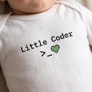 May include: A white short-sleeved baby bodysuit featuring the black text Little Coder followed by a pixelated greater-than sign, an underscore, and a small green pixelated heart symbol centered on the chest area of the garment.