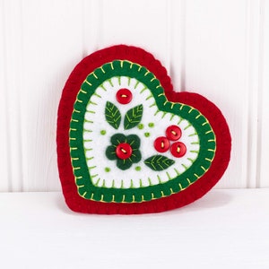 May include: A red felt heart ornament with green and white felt embroidery of flowers and berries. The ornament is decorated with red buttons.