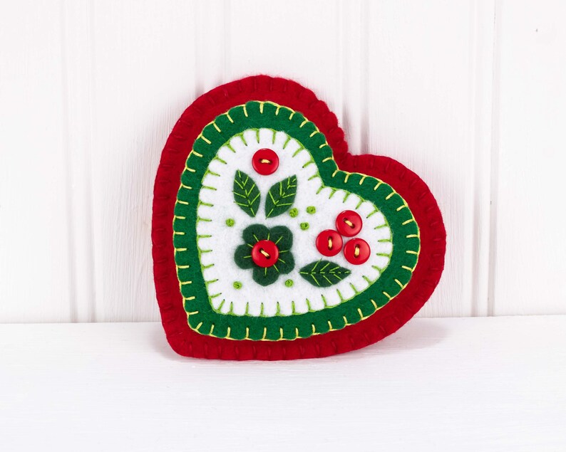 May include: A red felt heart ornament with green and white felt embroidery of flowers and berries. The ornament is decorated with red buttons.