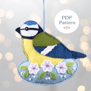 Op de afbeelding: Een vilten vogelornament in blauw, geel en groen, op een lichtblauwe basis met witte en paarse bloemen. Het heeft een blauw ophangkoord. De tekst "PDF Pattern" is zichtbaar.