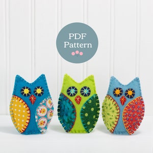 May include: Three felt owl ornaments, each with a different color and pattern. The owls are blue, green, and light blue with yellow, red, and white accents. The owls are all stitched together with a contrasting thread. The text "PDF Pattern" is visible in a circle above the owls.