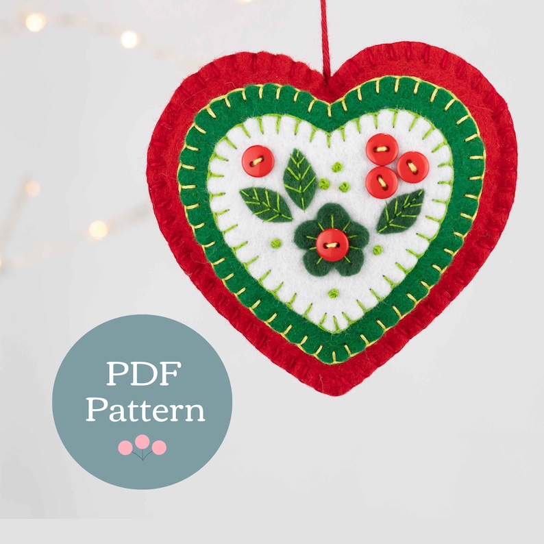 May include: A felt heart ornament with a green and red border and a white center. The center features a green flower with a red button center and green leaves with red button berries.