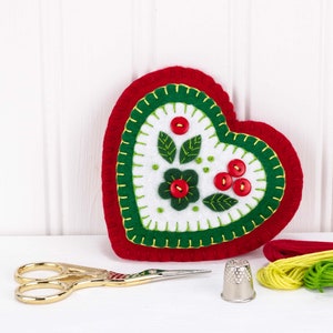 May include: A red felt heart with green and white embroidery of a flower and leaves. The heart is surrounded by red, yellow, and green embroidery floss.