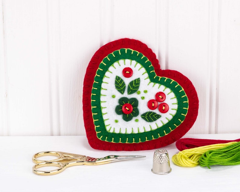 May include: A red felt heart with green and white embroidery of a flower and leaves. The heart is surrounded by red, yellow, and green embroidery floss.