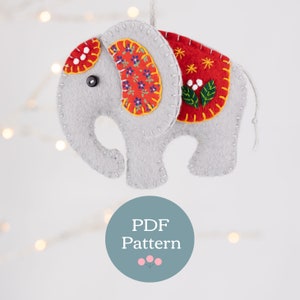 Può includere: Un ornamento di elefante in feltro grigio con tessuto a fantasia rossa, gialla e bianca sul dorso. L'ornamento ha un occhio a bottone nero e una corda bianca per appenderlo. Il testo "PDF Pattern" è su un cerchio blu sotto l'ornamento.