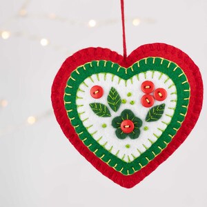 May include: A felt heart ornament with a green and red border and a white center. The center features a green flower with a red button center and green leaves with red button berries.
