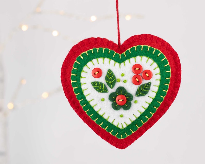 May include: A felt heart ornament with a green and red border and a white center. The center features a green flower with a red button center and green leaves with red button berries.
