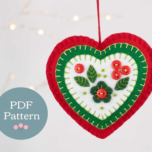 May include: A felt heart ornament with a red, green, and white design. The heart has a flower and leaves in the center, with red buttons and green stitching. The ornament is hanging from a string.
