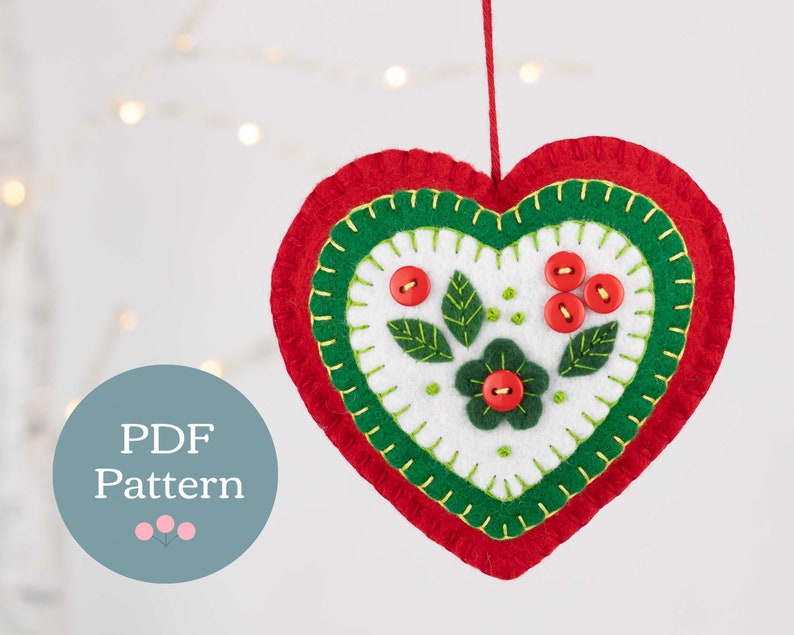 May include: A felt heart ornament with a red, green, and white design. The heart has a flower and leaves in the center, with red buttons and green stitching. The ornament is hanging from a string.