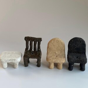 May include: Four miniature chairs and a table in a row. The table is white, the first chair is dark brown, the second is tan, and the last is black. Each piece has a textured, rough appearance.