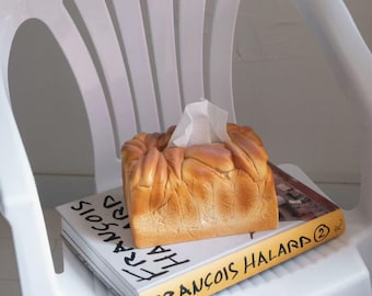 Puede incluir: Una caja de pañuelos con forma de pan sobre un libro titulado "FRANCOIS HALARD 2". El libro está sobre una silla de plástico blanca.
