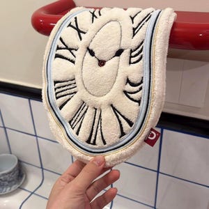 May include: A white, plush towel shaped like a melting clock face, featuring black Roman numerals and hands. The towel has a blue and gold border and is hanging on a red towel rack. A small red tag is visible.