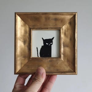 May include: A framed art print featuring a black silhouette of a grumpy cat. The cat has an angry expression with its ears perked up and tail raised. The artwork is set in a gold-colored, square frame.