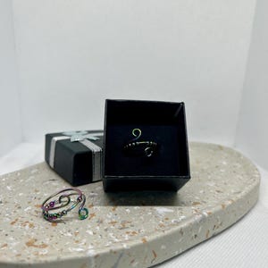Könnte beinhalten: Ein handgefertigter Drahtring mit schillernden Perlen und einem Spiraldesign liegt auf einer gesprenkelten grauen Oberfläche. Ein weiterer Ring wird in einer schwarzen Geschenkbox präsentiert. Eine schwarze Geschenkbox mit silbernem Band ist ebenfalls zu sehen.