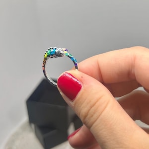 Op de afbeelding: Een zilveren ring met een kameleonontwerp met kleurrijke emaille details. Het lichaam van de kameleon is versierd met tinten blauw, groen, geel en paars. De ring wordt vastgehouden in een hand met rode nagellak.