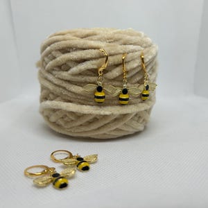 Könnte beinhalten: Goldfarbene Bienenohrringe mit schwarzen und gelben Details, präsentiert mit einem beigefarbenen Garnknäuel. Die Ohrringe haben einen Klappverschluss und zarte Flügeldesigns. Ein bezauberndes Accessoire.