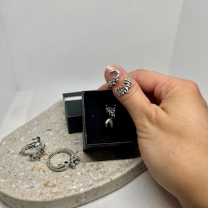Può includere: Una collezione di anelli argentati e una collana. Gli anelli presentano disegni intricati, tra cui un serpente, un artiglio e una forma curva. Un anello è su un dito. I gioielli sono esposti su un vassoio grigio chiaro maculato.
