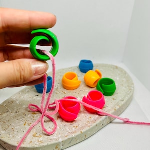 Könnte beinhalten: Eine Sammlung kleiner, farbenfroher, ringförmiger Bastelperlen in Grün, Blau, Orange und Pink. Ein rosafarbener Pfeifenreiniger ist durch die grüne Perle gefädelt. Die Perlen liegen auf einem ovalen Tablett mit Sprenkeln.