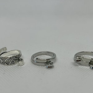 Op de afbeelding: Drie zilveren ringen worden getoond tegen een witte achtergrond. Eén ring is verstelbaar met een textuurontwerp en twee hebben een kleine bedel. De ringen zijn waarschijnlijk gemaakt van roestvrij staal of een vergelijkbaar metaal.