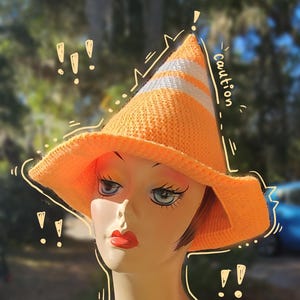 May include: A crocheted orange and white cone hat with a white stripe and the word "Caution" written on the side. The hat is worn by a mannequin with a neutral expression.