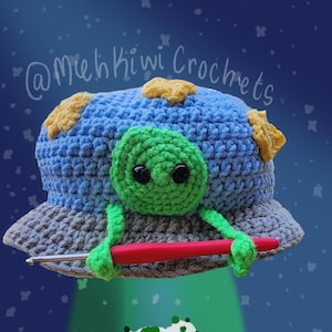 May include: A crocheted alien UFO with a green face and yellow stars on a blue background. The UFO is holding a red crochet hook and is beaming a green light down on a white and green crocheted cow.
