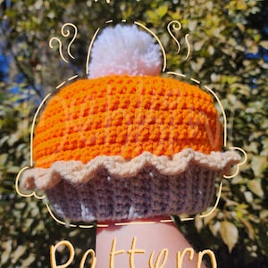 May include: A crocheted pumpkin pie hat with a white pom pom on top. The hat is orange and gray with a brown crust. The text "Pumpkin Pie Pattern" is written on the image. The text "@MehKiwiCrochets" is written at the bottom of the image.