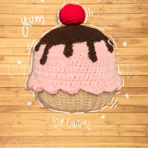 Puede incluir: Un gorro de cupcake tejido a crochet con glaseado rosa, un chorrito de chocolate marrón y una cereza roja en la parte superior. El gorro está sobre un fondo de madera con el texto "yum" y "so cute!" escrito en él.