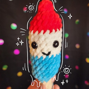 May include: A crocheted popsicle toy with a red, white, and blue color scheme. The popsicle has a smiling face with black eyes and a white mouth. The toy is held in a hand against a black background with colorful bokeh lights.