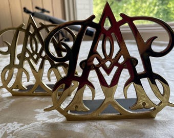 May include: Two gold-colored metal letter "A" decorative objects. The letters have an ornate, openwork design and are standing on a dark base. The objects are placed on a white patterned tablecloth.