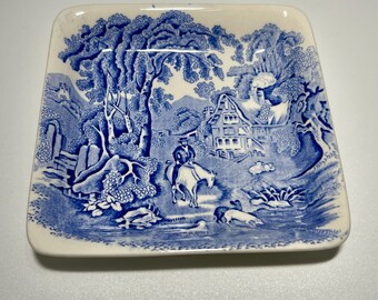 May include: Square ceramic dish with a blue and white pastoral scene. The design features a rider on horseback, a cottage, trees, and water. The dish has rounded corners and a cream-colored border.