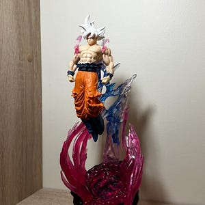 Op de afbeelding: Gedetailleerd beeldje van een personage met stekelig wit haar, een oranje broek en een gespierd lichaam. Het beeldje is omgeven door doorschijnende roze en blauwe energie-effecten en staat op een zwarte basis met gouden accenten. Thema: anime.