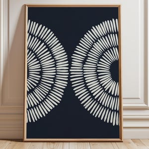Op de afbeelding: Abstracte kunstprint met twee witte, halfronde ontwerpen op een donker marineblauwe achtergrond. Het kunstwerk is ingelijst in een lichtbruine houten lijst en leunt tegen een witte muur en houten vloer.