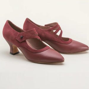 May include: A pair of dark red shoes featuring a pointed toe, a mid-height flared heel, and a dual-strap design across the instep. The shoes are crafted from a combination of smooth leather and suede textures, accented with a single dark button detail.